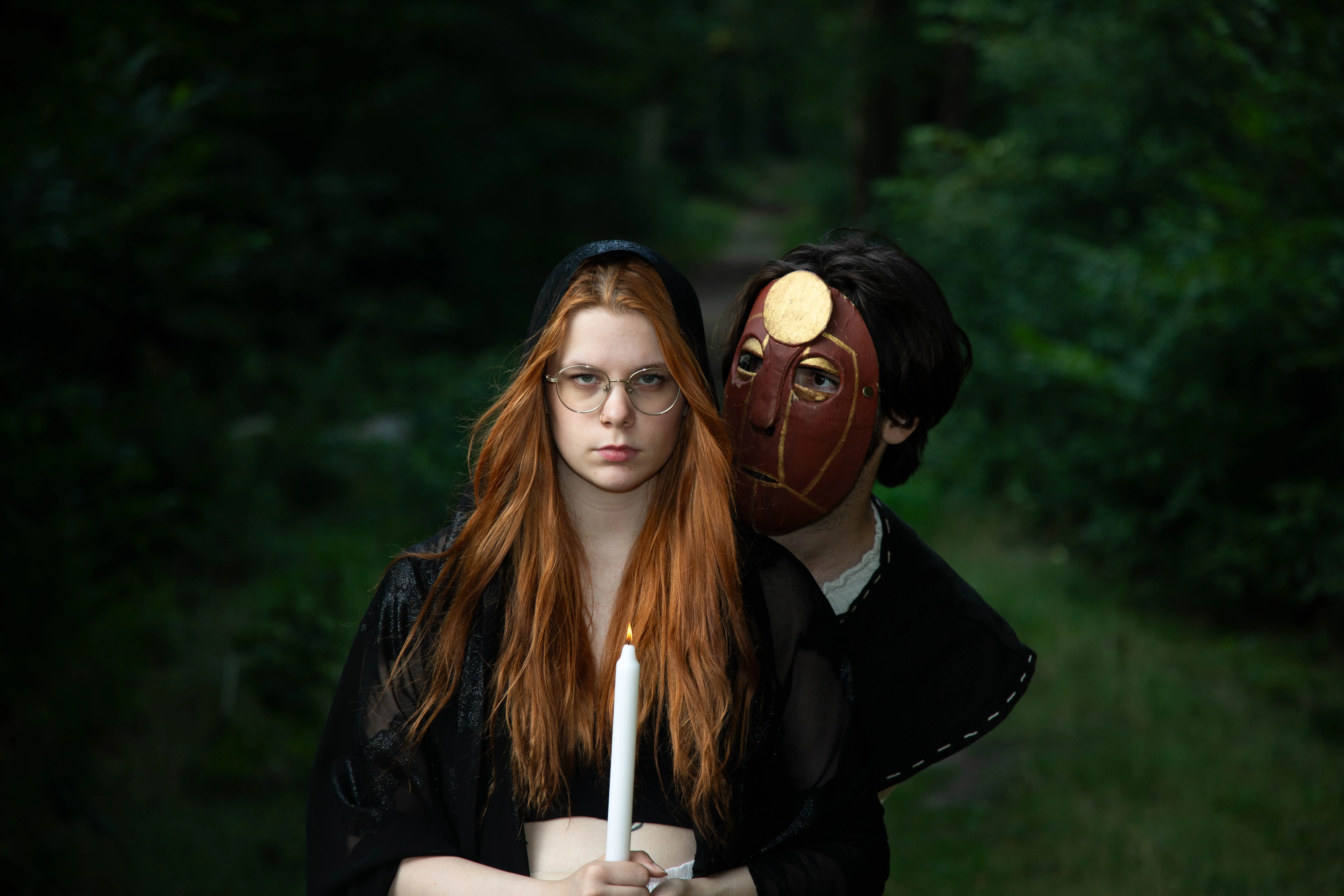 Een jonge vrouw met lang, rood haar en een kaars staat in een bos, terwijl een persoon met een maskerade achter haar staat.
