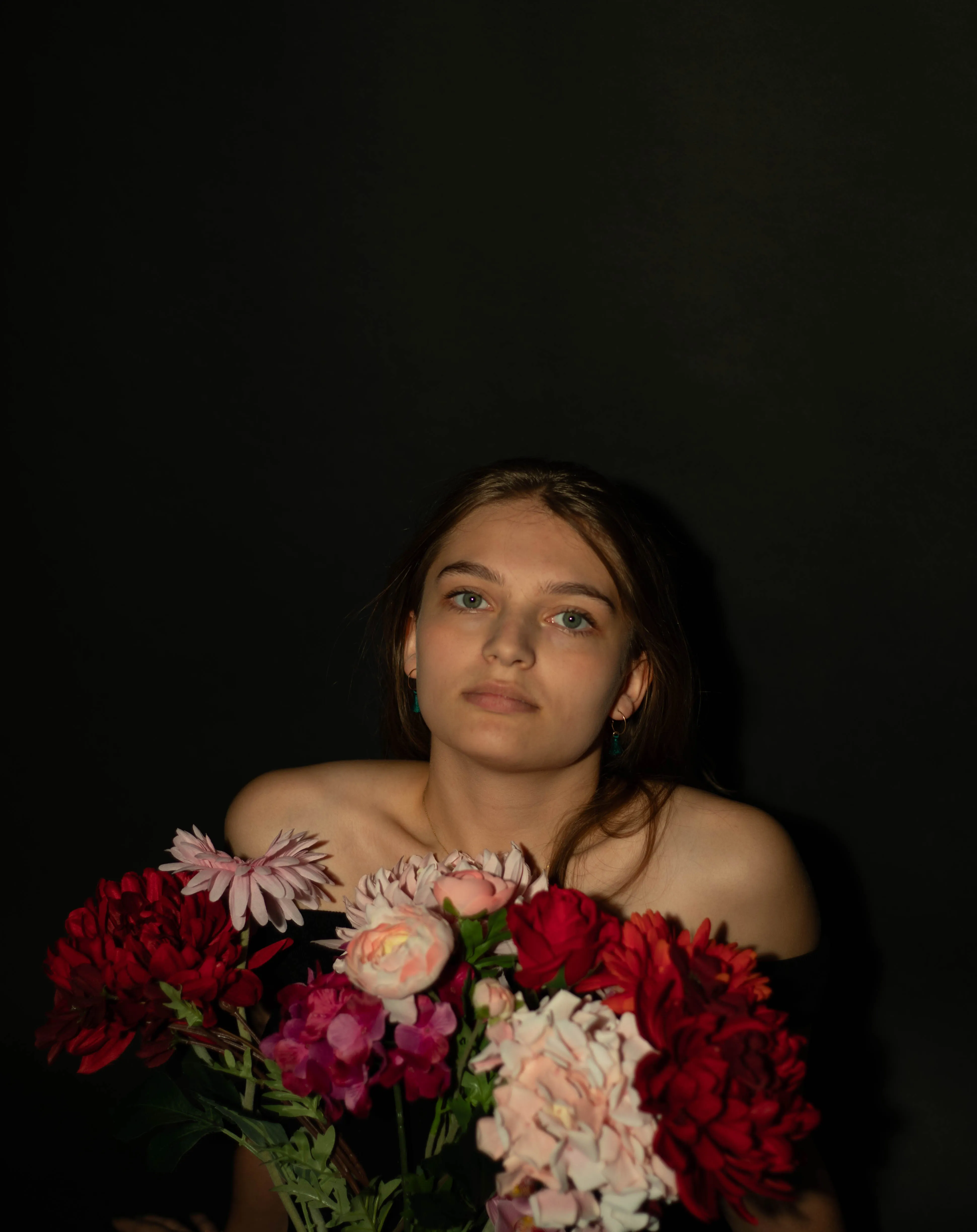 Een jonge vrouw met een neutrale uitstraling houdt een grote bos kleurrijke bloemen voor zich, tegen een donkere achtergrond.