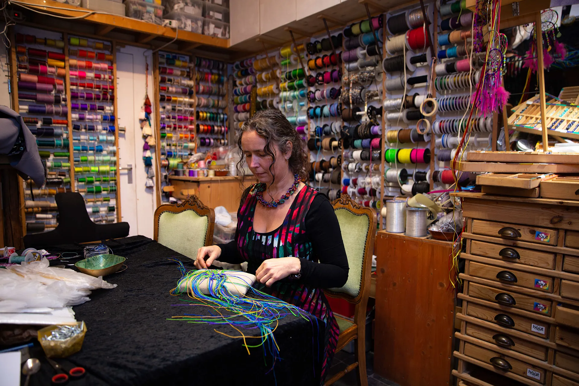 Een vrouw met krullend haar is bezig met het knopen van kleurrijke draden aan een tafel, omringd door een wand vol kleurrijke garens en materialen.
