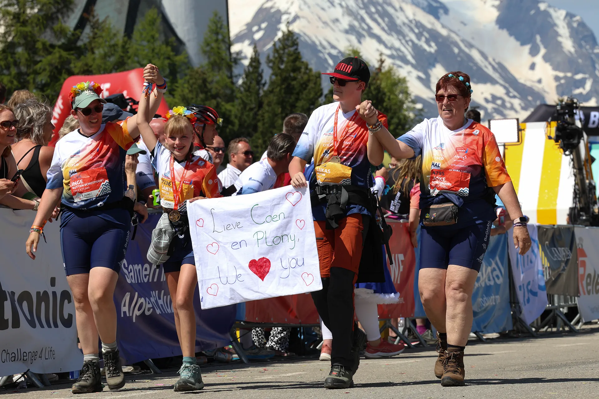 Een groep deelnemers aan een evenement houdt een banner vast met een lief bericht, terwijl ze vrolijk over de finishlijn lopen te midden van een feestelijke menigte en een bergachtige achtergrond.