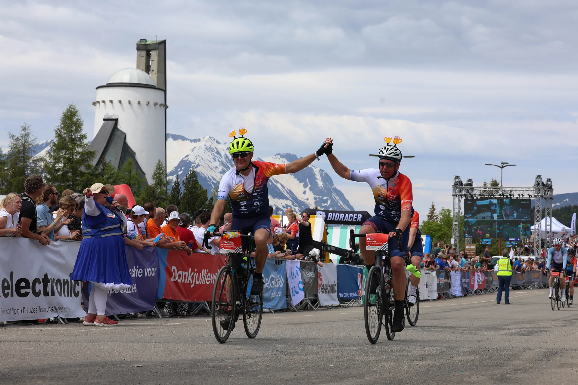 Twee fietsers vieren hun finish door elkaars handen vast te houden, terwijl een enthousiast publiek hen aanmoedigt en de bergen op de achtergrond zichtbaar zijn.