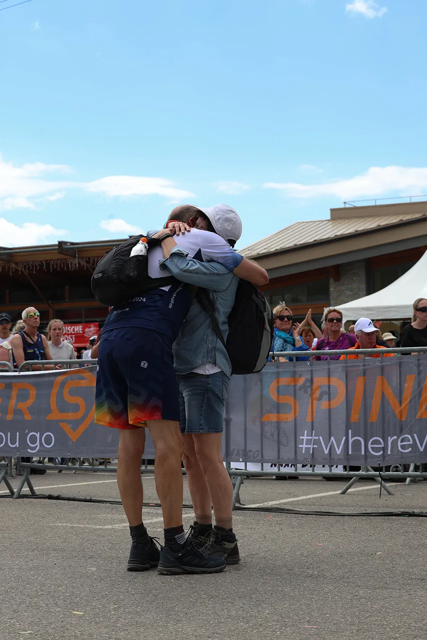 Twee mensen omhelzen elkaar hartelijk op een evenement, omringd door een menigte en een banner op de achtergrond.