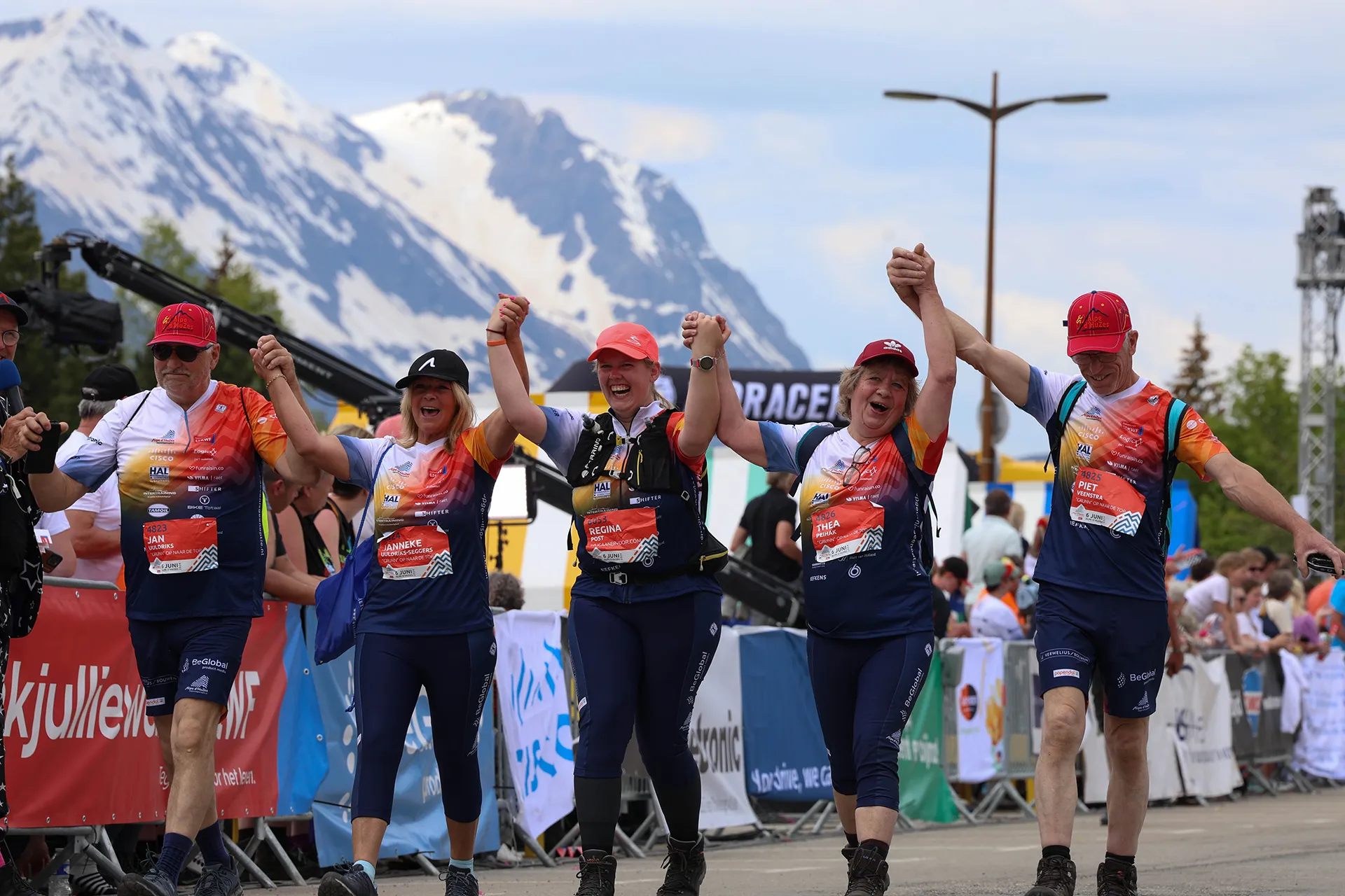 Een groep blijde deelnemers aan een race viert hun finish, terwijl ze elkaars handen vasthouden met een bergachtig landschap op de achtergrond.