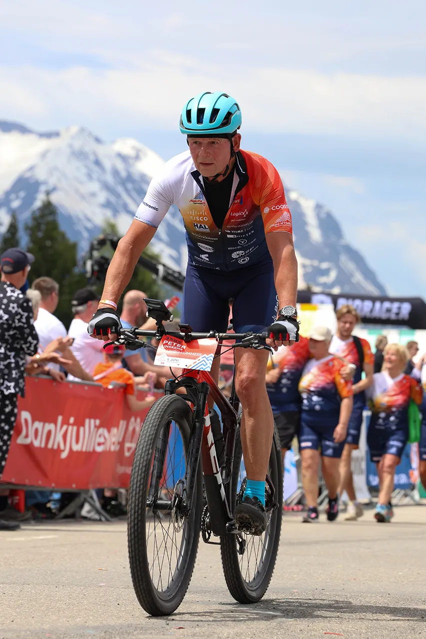 Een man in sportkleding fietst gefocust langs een menigte tijdens een race met bergen op de achtergrond.