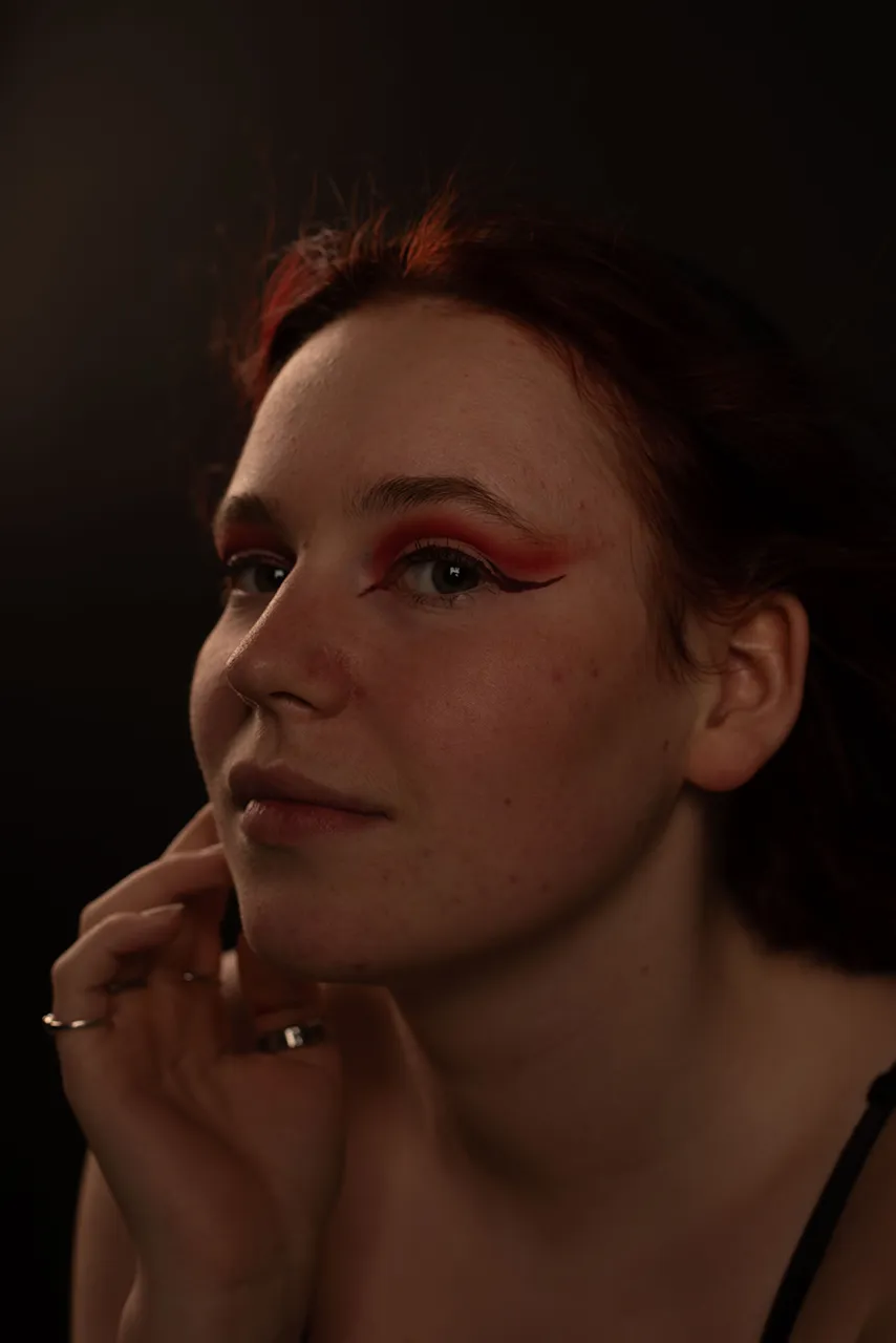 Een vrouw met een opvallende rode oogschaduw kijkt sereen naar de camera, met een hand tegen haar gezicht.