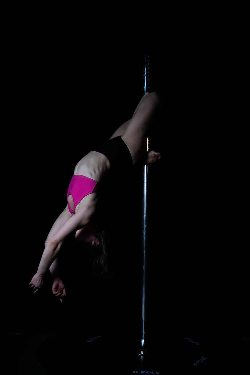 Een acrobatische danseres in een roze top hangt ondersteboven aan een paal tegen een donkere achtergrond.