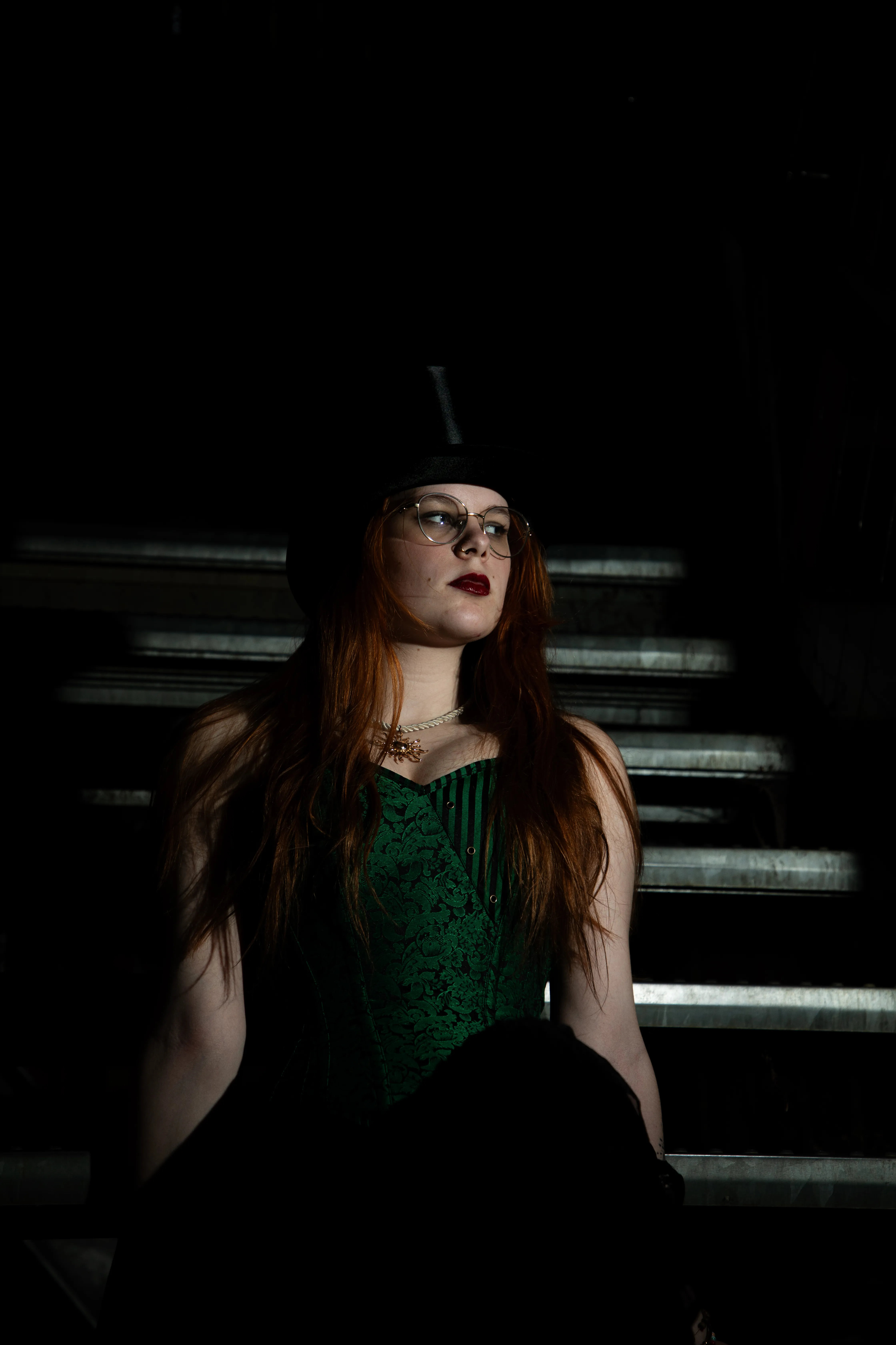 Een vrouw met lange, rode haren in een groene corset-top en hoge hoed, zittend op een trap in een donkere omgeving.
